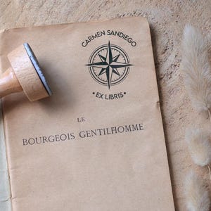 May include: A wooden stamp with a light-coloured handle rests on a vintage book page. The page is stamped with a compass rose design and the words "CARMEN SANDIEGO" and "EX LIBRIS". The book is titled "LE BOURGEOIS GENTILHOMME".