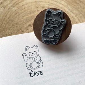 Könnte beinhalten: Ein Holzstempel mit einem lächelnden, winkenden Katzendesign und japanischem Text. Der Stempel ist auf eine Seite gedrückt und erzeugt eine Umrisszeichnung der Katze und den Namen "Élise" darunter. Der Hintergrund ist eine helle Holzoberfläche.