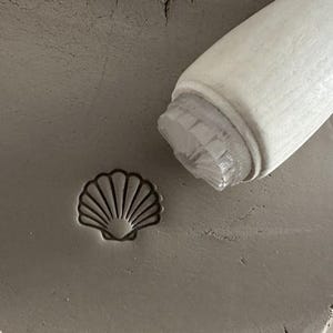 May include: A seashell-shaped stamp pressed into a grey clay surface. A white wooden handle tool is positioned nearby, suggesting the process of creating imprints. The image highlights the tools used in pottery or crafting.