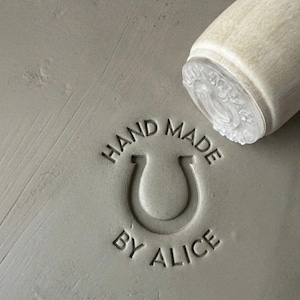 May include: A gray, textured surface with the words "HAND MADE BY ALICE" embossed around a horseshoe symbol. A wooden stamp with a clear, patterned base rests on the surface, suggesting a craft or handmade product.