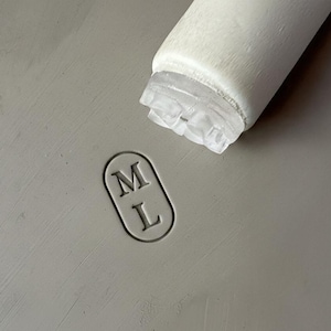 Peut inclure: Gros plan d'une surface grise avec un objet cylindrique blanc et une extrémité transparente et texturée. La surface présente un marquage ovale avec les lettres "M" et "L" à l'intérieur.