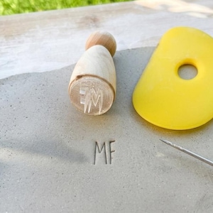 Op de afbeelding: Een lichtgrijze kleiplaat met de gestempelde letters MF, vergezeld van een houten stempel met hetzelfde MF-ontwerp, een geel flexibel niervormig pottenbakkersgereedschap van kunststof en een dunne metalen naald voor kleidetails.