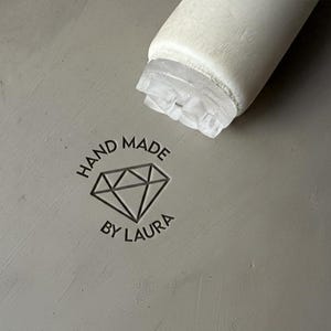 May include: Close-up of a gray surface with the text "HAND MADE BY LAURA" surrounding a diamond graphic. A white, cylindrical object with a clear, textured end is positioned at the top of the frame.
