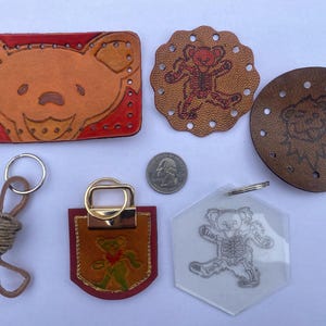 May include: A set of five leather keychains featuring a bear design. The keychains are in various colors and styles, including a brown leather keychain with a bear face, a red leather keychain with a bear face, a brown leather keychain with a bear outline, and a clear plastic keychain with a bear outline. The keychains are all attached to metal rings.
