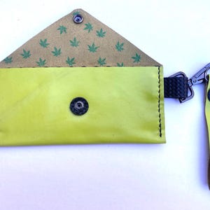 May include: A lime green leather pouch with a snap closure and black stitching. The interior flap features a pattern of green cannabis leaves. A black wrist strap and silver-tone clasp are attached.