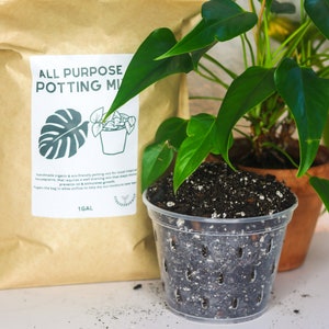 May include: A clear plastic pot filled with potting mix sits next to a brown paper bag of potting mix. The bag has a white label with the text "ALL PURPOSE POTTING MIX" and a graphic of a plant. A potted plant with green leaves is in the background.