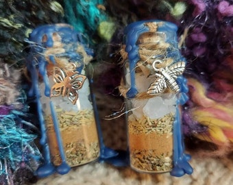 May include: Two small glass bottles filled with herbs and crystals, sealed with blue wax and adorned with silver charms. The bottles are decorated with twine and a butterfly and bee charm. The background is a colorful, textured fabric.
