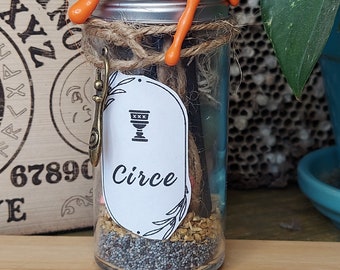 May include: A glass jar with a silver lid, twine, and an orange wax seal. The jar contains seeds and sticks, with a label that reads "Circe" and a chalice symbol. A decorative charm hangs from the twine.