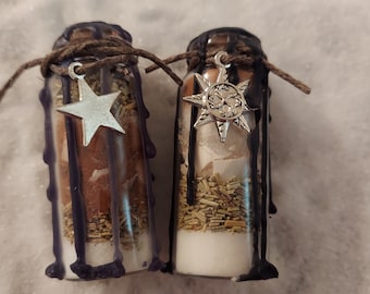 May include: Two small glass bottles filled with herbs and powders, sealed with dark wax and twine. Each bottle has a silver charm, one a star and the other a sunburst. The bottles are on a white, textured surface.