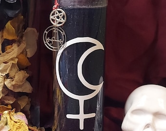 Puede incluir: Una vela negra alta en un frasco de vidrio transparente, adornada con una luna creciente blanca y un símbolo femenino. Un dije plateado con un pentagrama y otros símbolos cuelga de la parte superior. La vela está decorada con hierbas secas y atada con hilo rojo.