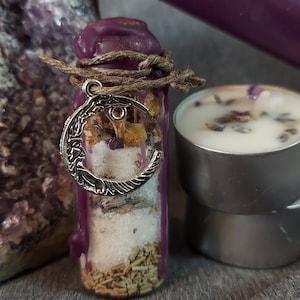May include: A purple-wax-sealed glass bottle filled with herbs and crystals, adorned with a silver crescent moon charm. Two tea light candles with lavender and a purple candle are also present. The items are arranged on a gray surface.