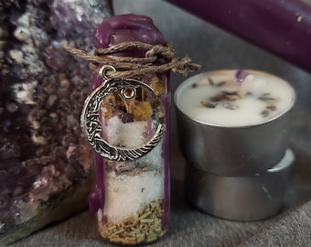 May include: A purple-wax-sealed glass bottle filled with herbs and crystals, adorned with a silver crescent moon charm. Two tea light candles with lavender and a purple candle are also present. The items are arranged on a gray surface.