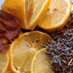May include: A collection of dried botanical elements including bright yellow lemon slices, deep maroon dried rose petals, and a mound of small, dried purple lavender buds arranged together to create a textured, colorful, and aromatic display.