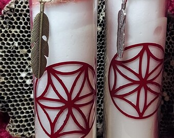 May include: Two tall, cylindrical candles with a white wax base and a pink top. Each candle has a red geometric design and a decorative feather charm. The candles are in glass jars, adorned with twine and small embellishments.