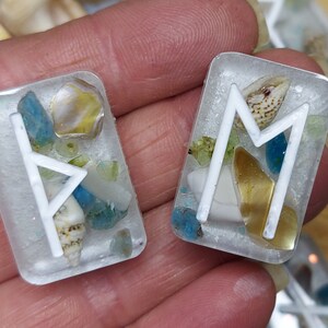 May include: Two rectangular resin pendants with white rune symbols. The pendants are filled with small seashells, blue and yellow stones, and other decorative elements. The background is a light color.
