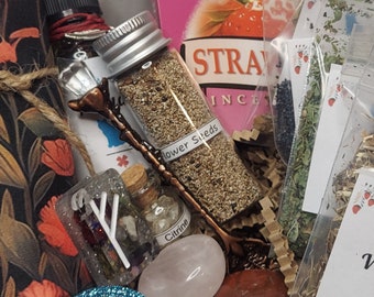 May include: A close-up of a witchy gift box filled with various items, including a small bottle of flower seeds, a clear glass bottle with a white label that says "Citrine", a pink rose quartz, a brown stone, a small glass bottle with a red liquid, a small wooden spoon, a small piece of paper with a white rune symbol, and a small bag of dried herbs. The box is filled with brown packing peanuts.