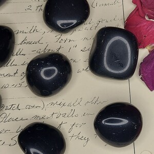May include: A collection of seven smooth, black, polished stones with a slight sheen. The stones are arranged on a piece of paper with handwritten text.