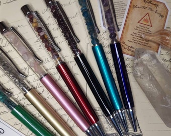 May include: A collection of eight pens with clear barrels filled with various colorful stones and crystals. The pens have different colored metal barrels, including gold, silver, pink, red, black, teal, and blue.