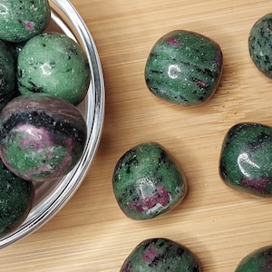 May include: A collection of tumbled ruby in zoisite stones. The stones are a mix of green and black zoisite with patches of red ruby. Some stones are in a clear glass bowl, while others are scattered on a wooden surface.