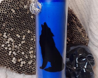 May include: A blue candle with a black silhouette of a howling wolf. The candle has a silver charm and is decorated with a black string and small sticks. The candle is in a glass jar.