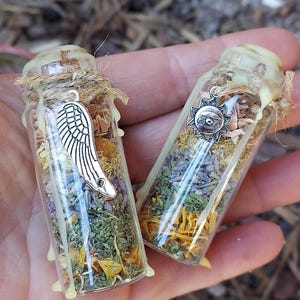 May include: Two small glass bottles filled with dried herbs and flowers. Each bottle is adorned with a silver charm, one a wing and the other a sun. The bottles are sealed with a light-colored wax and twine.