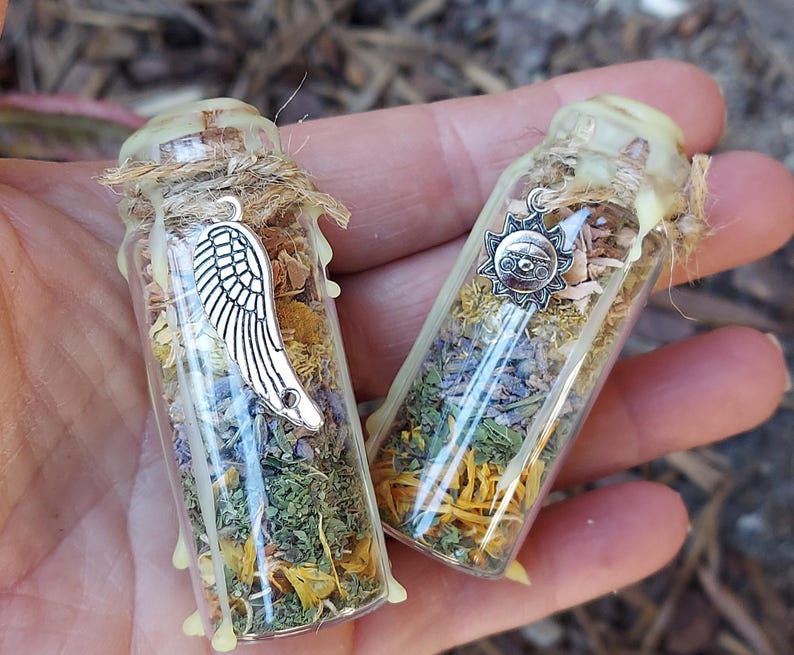 May include: Two small glass bottles filled with dried herbs and flowers. Each bottle is adorned with a silver charm, one a wing and the other a sun. The bottles are sealed with a light-colored wax and twine.