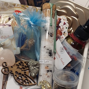 May include: A collection of self-care items, including a glass tube of bath salts, a small bottle of oil, and a blue organza bag filled with crystals. Other items include a wooden cutout, a heart-shaped stone, and a small jar with a silver lid.