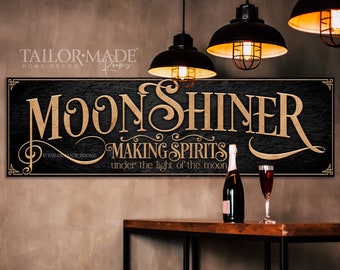 May include: Black wooden sign with gold lettering that reads "Moonshiner Making Spirits under the light of the moon". The sign is hanging on a wall with two black pendant lights above it.
