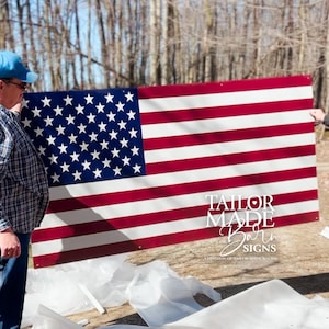 May include: A large American flag with red and white stripes and a blue field of white stars. The flag is being held up outdoors. The text "TAILOR MADE Barn SIGNS" is visible in the lower right corner.