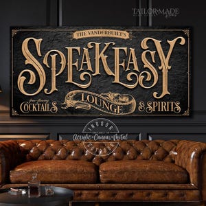 May include: A rectangular wall sign with a black textured background and gold lettering. The text reads THE VANDERBUILT'S SPEAKEASY LOUNGE & SPIRITS with free-flowing COCKTAILS. The design features decorative scrollwork and borders in a gold finish.