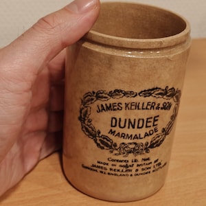 May include: A vintage, cylindrical stoneware jar with a light brown hue. The jar features a black and white label with the text "JAMES KEILLER & SON DUNDEE MARMALADE". The jar is held by a hand.