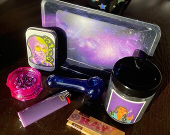 May include: A galaxy-themed rolling tray with a purple and blue design, a small grinder with a pink lid, a blue glass pipe, a purple lighter, a pack of RAW Classic rolling papers, and a small tin with a cartoon image of a woman with a cigarette.