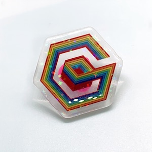 May include: A clear, hexagonal pin with a rainbow-colored geometric design. The pin features a series of nested shapes in vibrant colors, including red, orange, yellow, green, blue, and purple. The pin has a holographic, glittery finish.