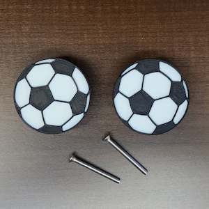 May include: Two soccer ball-shaped cabinet knobs with black and white panels. Two silver screws are positioned below the knobs. The knobs are round and have a 3D printed appearance. The background is a dark brown wooden surface.