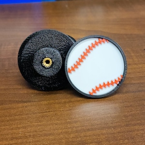 Peut inclure: Deux objets ronds, noirs, imprimés en 3D. Un objet est une balle de baseball avec des coutures rouges. L'autre objet a un petit trou au centre.