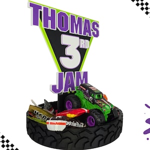 May include: A birthday cake topper with a green and black monster truck on a tyre base. The topper includes a sign with the name "THOMAS" and the number "3" in purple and white. Candy is also included.