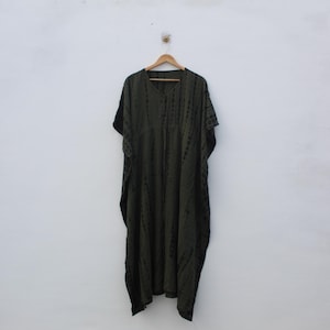 Może przedstawiać: Oliwkowozielona sukienka kaftan z dekoltem w serek i krótkimi rękawami, wisząca na drewnianym wieszaku. Sukienka ma wzór tie-dye w ciemniejszych odcieniach zieleni i czerni, tworząc teksturowany wygląd. Sukienka jest wykonana z lekkiego materiału.