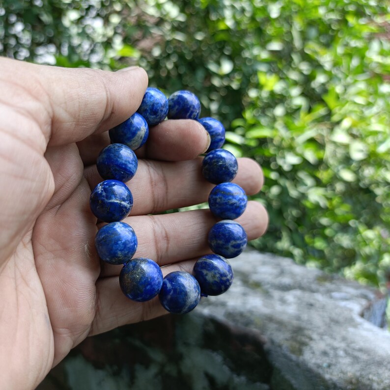 Puede incluir: Una pulsera de cuentas azul oscuro con apariencia de piedra natural. La pulsera est&aacute; compuesta por cuentas redondas con una textura veteada, unidas por una banda el&aacute;stica. La pulsera se sostiene en una mano sobre un fondo verde borroso.