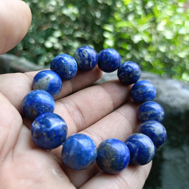 Puede incluir: Un brazalete con cuentas esf&eacute;ricas de lapisl&aacute;zuli azul profundo, con inclusiones doradas y azul claro. Las cuentas est&aacute;n ensartadas para formar un accesorio port&aacute;til. El brazalete se sostiene en una mano.