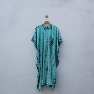 May include: A turquoise tie-dye caftan dress with a V-neck and short sleeves hangs on a wooden hanger. The dress features vertical dark blue stripes and a loose, flowing silhouette. The background is a plain white wall.