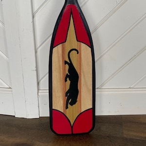 May include: A wooden paddle with a black panther silhouette painted on the front. The paddle is painted with red and black stripes and has a red and black design at the top.