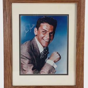 May include: Framed portrait of a smiling man in a suit and bow tie, set against a blue backdrop. The portrait is signed. The frame is made of wood.
