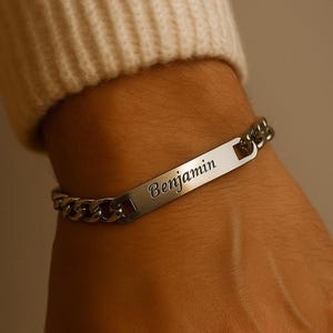 Peut inclure: Un bracelet argenté avec une plaque nominative rectangulaire gravée du nom "Benjamin". Le bracelet présente un motif de chaîne gourmette. L'image montre le bracelet au poignet d'une personne, avec un pull crème en arrière-plan.