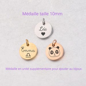 Può includere: Tre piccoli ciondoli rotondi con un anello per appenderli. Uno è argentato con il nome "Léa" e un cuore, un altro è dorato con il nome "Emma" e un simbolo zodiacale, e il terzo è oro rosa con una faccia di panda.