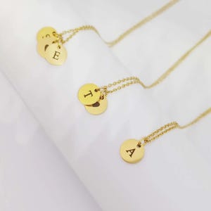 Puede incluir: Collares dorados con delicadas cadenas. Cada collar presenta un colgante redondo con una inicial grabada. Los colgantes están en un grupo de dos o tres. El fondo es blanco.