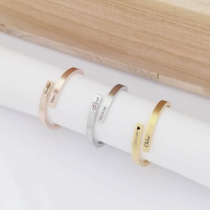 Peut inclure: Trois bracelets manchettes personnalisés en or rose, argent et or. Chaque bracelet porte un nom et/ou une date gravés. Les bracelets sont présentés sur une surface cylindrique blanche sur un fond en bois clair.