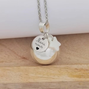 Peut inclure: Collier argenté avec un pendentif sphérique, un petit charme rond avec l'inscription "Mini nous" et un cœur, et un charme en forme d'étoile. Le collier est suspendu à une chaîne argentée. Le fond est une surface en bois clair.