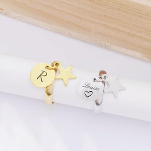 Könnte beinhalten: Zwei Ringe, einer goldfarben und einer silberfarben, jeweils mit einem Anhänger. Der goldfarbene Ring hat einen runden Anhänger mit dem Buchstaben "R" und einem Sternanhänger. Der silberfarbene Ring hat einen runden Anhänger mit dem Namen "Louise" und einem Herzsymbol sowie einen Sternanhänger.
