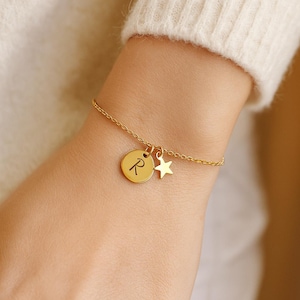Könnte beinhalten: Goldfarbenes Armband mit feiner Kette. Es hat einen runden Anhänger mit dem Buchstaben "R" und einen kleinen Sternanhänger. Das Armband wird am Handgelenk getragen, vor einem cremefarbenen Pullover.