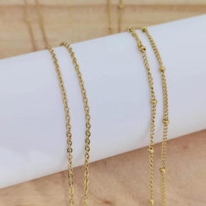 Peut inclure: Gros plan de plusieurs colliers de couleur or. Les chaînes ont différents designs, dont une chaîne à maillons classique et une chaîne avec de petits accents de boules. Les colliers sont présentés sur une surface blanche avec un fond en bois.